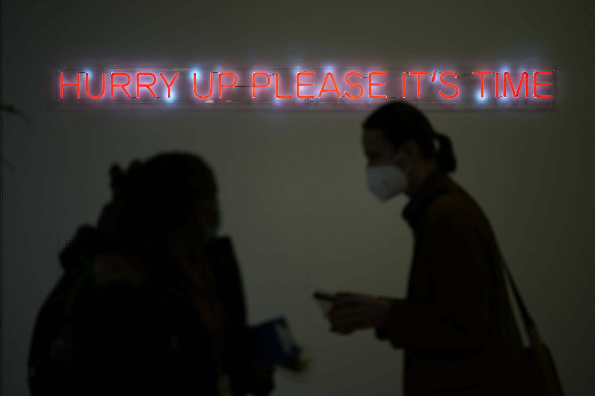 silhouettes of people walk past a neon sign that says hurry up please it